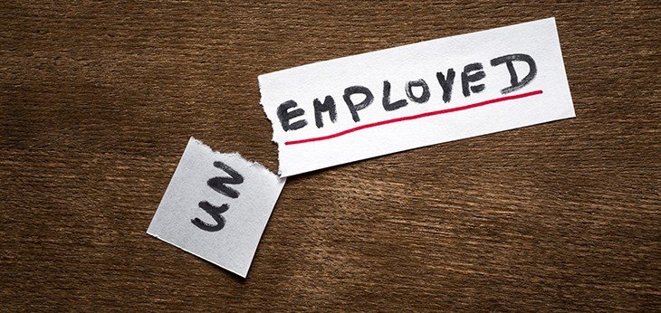 Slim piece of paper on a dark wooden background. On the paper is the word unemployed written in heavy dark marker and underlined in red. The paper has been torn at after the letters 'un' and this piece is on an angle.