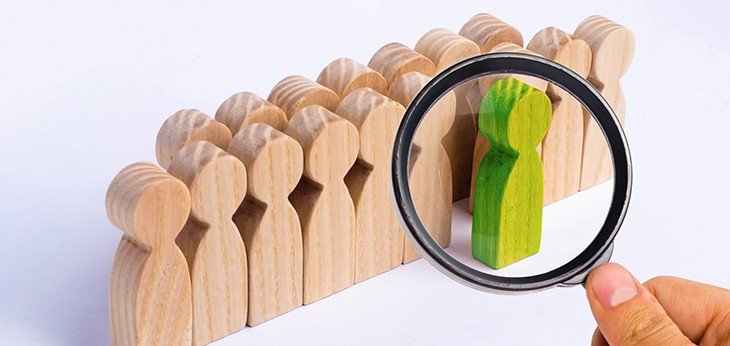 Wooden figures representing people with one green in front ringed by a magnifying glass