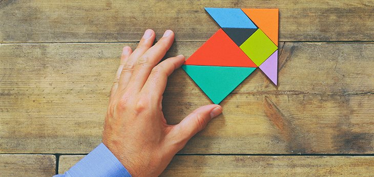 a mans hand pushing a colourful arrow made up of puzzle pieces on a wooden table