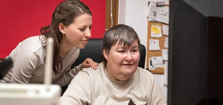 Woman sitting in front of a computer with her carer standing behind assisting